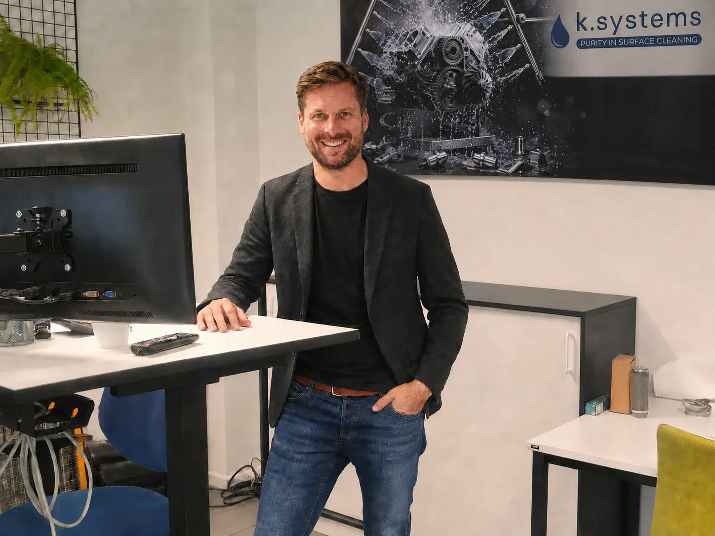 Patrick Kölle im dunklen Sakko am Arbeitsplatz vor dem k.systems-Wandmotiv